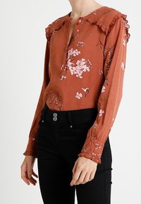 Rostfärgad blus med blommönster, volangdetaljer på axlarna, knäppning fram och långa ärmar. Matchad med svarta höga byxor.