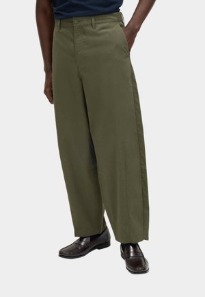 Persoon draagt wijde olijfgroene broek, zwarte sokken, donkerbruine leren loafers en een donkerblauw overhemd in de broek gestopt.