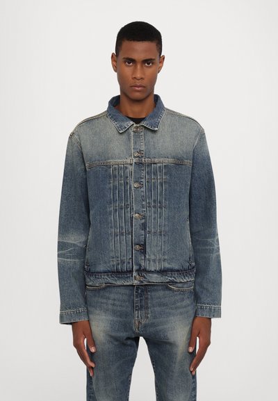 Veste en denim avec une fermeture à bouton, un col à pointes et un côtelage vertical sur la poitrine. La couleur est un bleu moyen avec une légère décoloration.