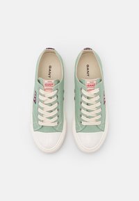 Světlé zelené plátěné tenisky s bílými gumovými špičkami, béžovými tkaničkami a červenou a bílou logo nášivkou na boku.