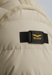 Veste rembourrée beige avec une texture lisse, dotée d'une sangle en tissu noir et d'un logo détaillé avec du texte jaune et des graphiques d'ailes.