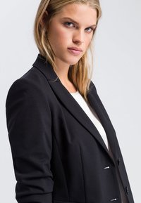 Black blazer with a tailored fit, single-breasted design, peaked lapels, and two buttons. Smooth fabric with structured shoulders.
