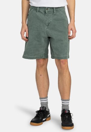 Grønne bomuldsshorts, knælange, med en lige snitdesign, forlommer og en syet kant. Båret med sorte sneakers og gråstribede sokker.