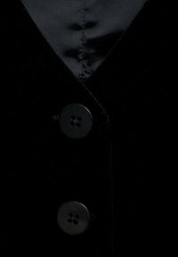 Černá sametová vesta s výstřihem do V, zdobená třemi lesklými černými knoflíky a hladkou podšívkou. Viditelné jemné prošívání.