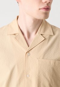 Beige kurzärmliges Hemd mit Knopfleiste und strukturierter Oberfläche, spitzem Kragen und einer Brusttasche. Mit weißen Knöpfen.