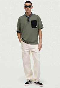 T-shirt verde-azeitona com um bolso preto, calças em tom creme e ténis pretos. Os acessórios incluem um lenço estampado e óculos de sol.