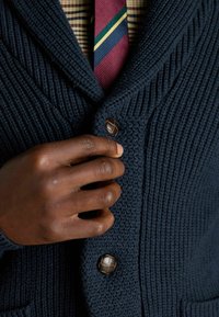 Marineblauer Strickcardigan mit einem Schalkragen, aus strukturiertem Stoff, großen braunen Knopfakzenten und einem sichtbaren karierten Hemd darunter.