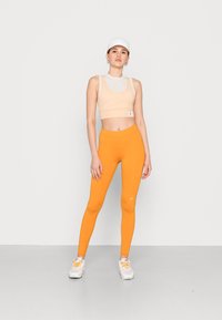 Crop top sportivo beige con dettagli in rete, leggings arancioni e sneakers bianche con accenti arancioni. Design semplice, silhouette aderente.