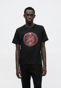 Mustast puuvillast T-särk, millel on punase värvusega ümmargune graafika tegelasega, kellel on käed risti. Lühikesed varrukad ja ümara kaelusega disain.