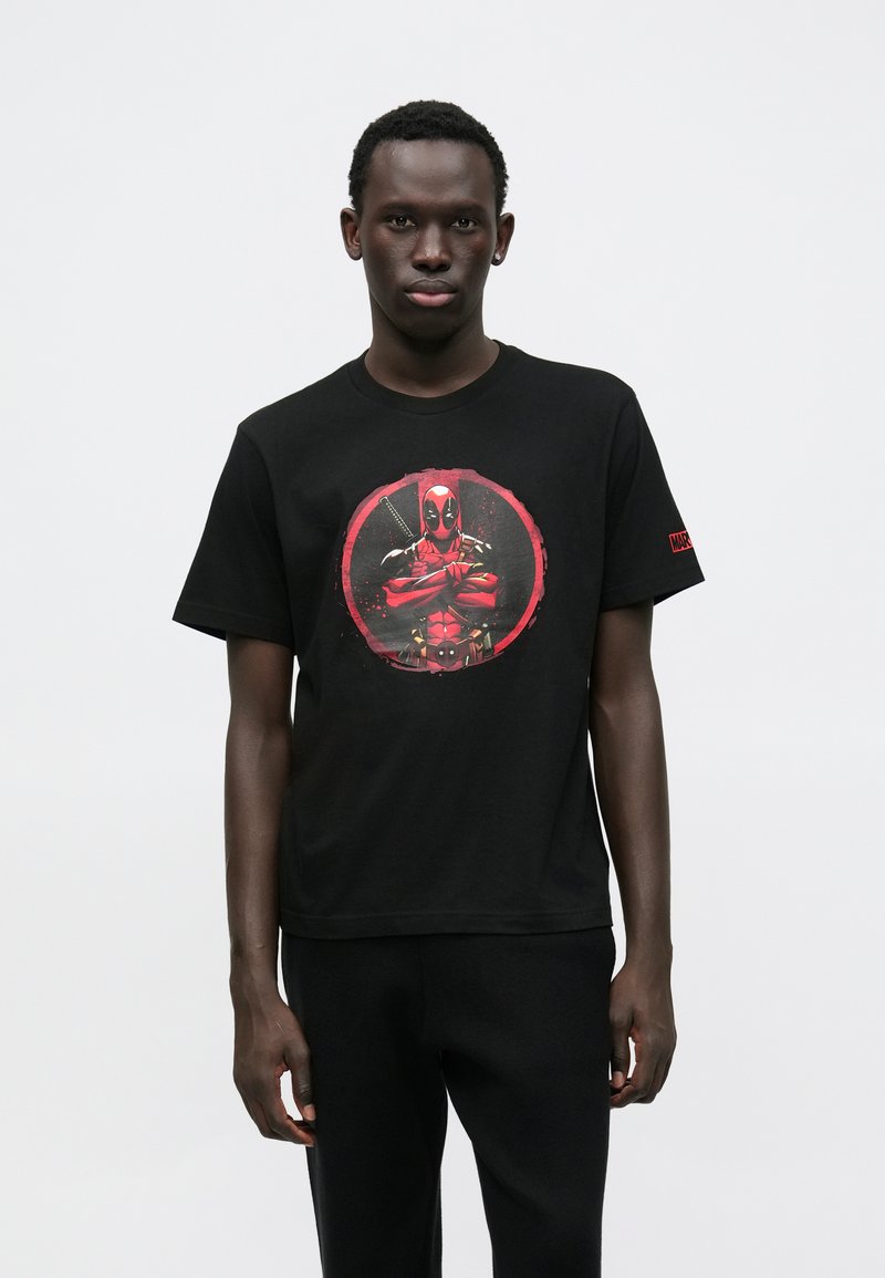 Mustast puuvillast T-särk, millel on punase värvusega ümmargune graafika tegelasega, kellel on käed risti. Lühikesed varrukad ja ümara kaelusega disain.