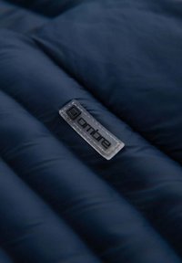 Marineblaue Steppjacke mit gestepptem Muster; verfügt über ein transparentes Etikett mit dem angebrachtem "ombre"-Logo.