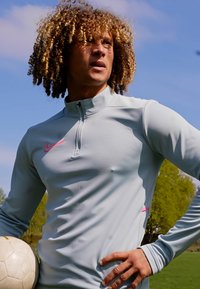 Lyseblå langærmet sportstop med hætte og halvlukning. Har et pink Nike-logo på venstre bryst. Glat stof, tætsiddende form.