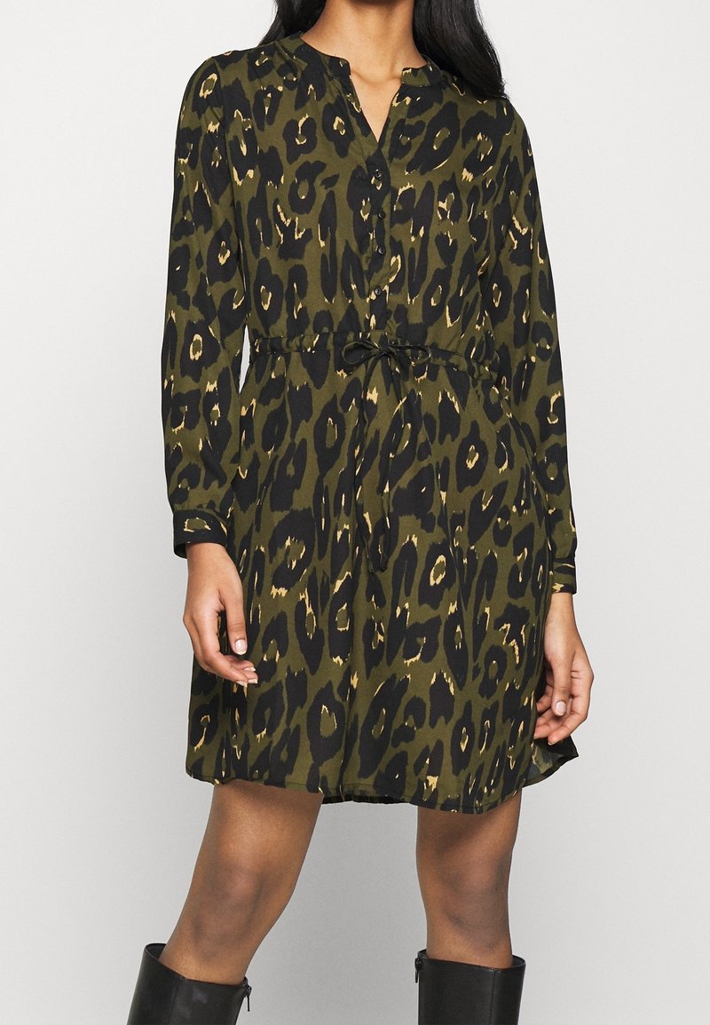 Femme portant une robe mi-longue vert olive et noir à imprimé léopard, manches longues, avec des boutons sur le devant, associée à des bottes noires.