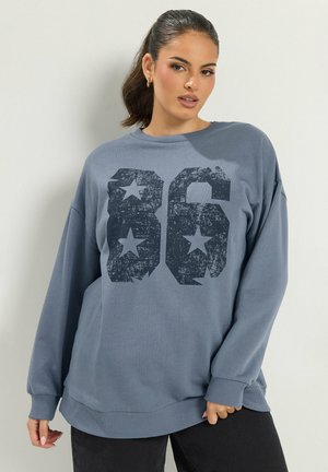 Jeune femme aux cheveux tirés en arrière porte un sweat-shirt bleu ample avec le numéro 86 usé et des étoiles, posant devant un mur clair et uni.