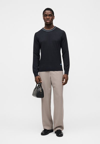 Pull noir à manches longues avec un col rayé bleu, associé à un pantalon beige clair sur mesure et un petit sac à main noir.