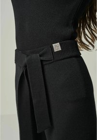 Schwarzes geripptes Kleid mit einem gebundenen Taillengürtel und einem kleinen silbernen Logo-Tag. Der Stoff hat eine weiche Textur und ein figurbetontes Design.