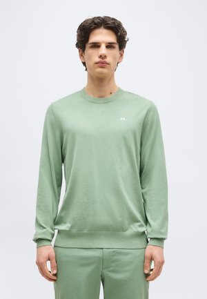 Νεαρός άνδρας με κοντά σκούρα μαλλιά, φορώντας ανοιχτό πράσινο πουλόβερ με στρογγυλή λαιμόκοψη και αντίστοιχο παντελόνι, στέκεται μπροστά σε ένα απλό λευκό φόντο.