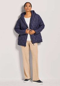 Marineblau gesteppte Kapuzenjacke mit Reißverschluss, langen Ärmeln und lockerer Passform, kombiniert mit hellbeigen Hosen und dunklen Sneakers.