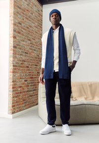 Dunkelblauer Strickmütze, gerippter Schal, cremfarbener ärmelloser Pullover über einem weißen Hemd, dunkle Hose und weiße Turnschuhe. Strukturierte Backsteinwand im Hintergrund.