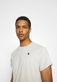 Beige bomull t-skjorte med korte rullede ermer, med en liten svart logo på venstre bryst, myk tekstur, og en klassisk rund hals.