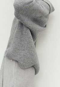 Personne portant un pull gris ample à côtes et un pantalon gris clair, les bras levés, se tenant devant un fond blanc uni.