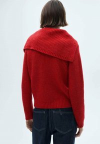 Pull en tricot rouge avec un large col texturé et une taille côtelée. Le dos présente une coupe décontractée et est associé à un jean bleu foncé.
