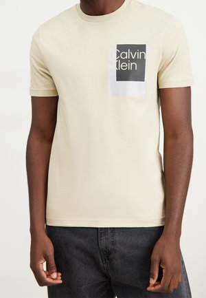 Muž nosící béžové tričko s krátkým rukávem Calvin Klein s černobílým logem na hrudi, spojené s tmavě šedými džíny.