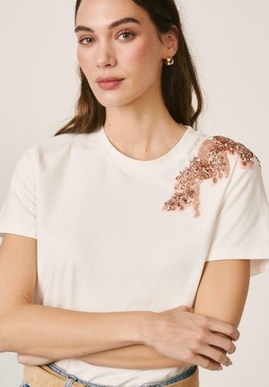 Mujer de piel clara y cabello castaño lleva una blusa blanca de manga corta con un adorno floral de cuentas rosas en el hombro, con los brazos cruzados.