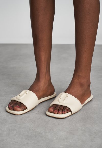 Beige slide sandals made of soft material, featuring a raised decorative buckle on the strap and a square toe design.