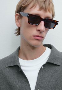  Schildpad zonnebril met donkere glazen, rechthoekige vorm; model draagt een grijze cardigan over een wit T-shirt met ronde nek. Vloeiende texturen, minimale details.