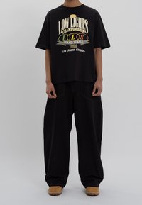 Černé oversized tričko s grafickým textem, v kombinaci s volnými černými kalhotami a béžovou obuví. Hladký, ležérní design s minimální texturou.