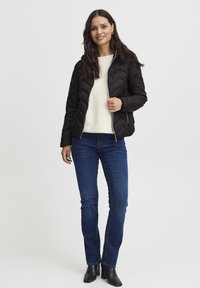 Črna prešita jakna z zadrgo in vzorcem ribje kosti, v kombinaciji s krem puloverjem in temnordečimi kavbojkami, nošena s črnimi gležnjaki.