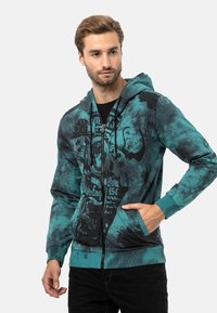 Kapuzensweatshirt in Türkis und Schwarz, mit einem Reißverschluss, Fronttaschen und grafischen Drucken. Hergestellt aus weichem Material mit einer strukturierten Oberfläche.