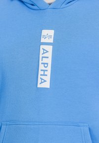 Modra kapuca iz mehkega materiala, ki ima beli logotip s tiskom "ALPHA" in grafične detajle na sprednji strani. Vidna je sprednja žepa.
