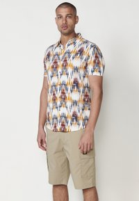 Chemise à manches courtes avec un motif géométrique coloré en orange, jaune et bleu sur fond blanc. Associée à un short cargo beige.