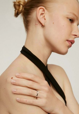 Anillo de plata con una piedra preciosa central, llevado en una mano que descansa sobre un hombro desnudo. El modelo lleva una blusa halter negra, lo que destaca prominentemente el anillo.