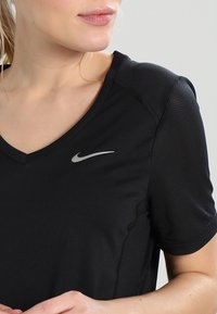 Camiseta deportiva negra con diseño de cuello en V, confeccionada con una tela suave; presenta un área texturizada en los hombros y un logo reflectante de Nike.