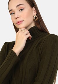 Olijfgroen gebreid trui met een hoge kraag en een gestructureerd patroon. Model draagt gouden hoepeloorbellen en ringen, en maakt handgebaren.