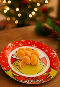 Piatto rosso con un design del Grinch in stile cartone animato e la scritta "MERRY GRINCHMAS". Il piatto contiene segmenti di mandarino arancioni disposti ordinatamente.