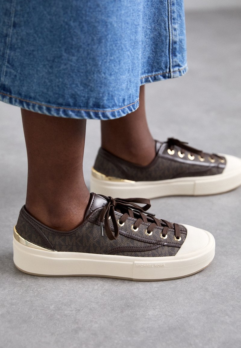 Personne portant des baskets basses Michael Kors marron avec des semelles blanches et des accents dorés, associées à une jupe en jean bleue sur un sol gris.