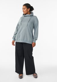 Felpa grigia con scollo a cowl, maniche lunghe, vestibilità comoda e dettagli con coulisse. Abbinata a pantaloni neri a gamba larga e scarpe con motivo.