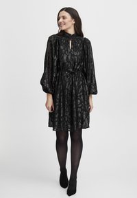 Rochie neagră cu guler înalt, detaliu în formă de cheie și mâneci puf lungi. Prezintă un model texturat și o curea la talie. Material neted.