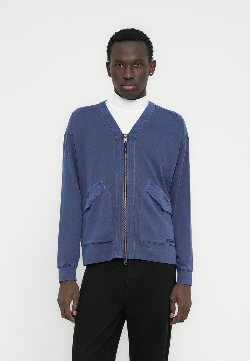 Μπλε μπουφάν με φερμουάρ και V-λαιμόκοψη, μακριά μανίκια, δύο μπροστινές τσέπες και υφή με ανάγλυφη επιφάνεια. Φορεμένο πάνω από λευκό ζιβάγκο και συνδυασμένο με σκούρο παντελόνι.