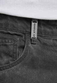 Detailní záběr na pas tmavě modrých džín s kovovým štítkem označeným "2Y STUDIOS" a bílým žebrovaným tričkem zasunutým uvnitř.