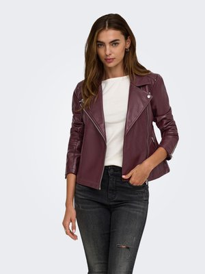Donna con capelli lunghi e castani che indossa una giacca di pelle color bordeaux, un top bianco e jeans neri strappati, in piedi con una mano in tasca.