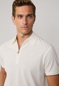 Biely polo s zipsom pri golieri, s jemným logom na hrudi a kontrastným lemovaním. Vyrobené z hladkej látky s uvoľneným strihom.