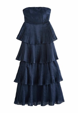 Kobieta w granatowej, bezramkowej, warstwowej plisowanej sukni, trzymająca małą złotą torebkę, stojąca na tle jasnoszarej ściany.