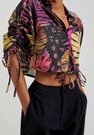 Blouse courte boutonnée avec imprimé de feuilles tropicales violettes, jaunes et roses, manches froncées et nœud à l'avant, portée avec un pantalon noir et les mains dans les poches.