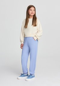 Felpa corta di colore crema e pantaloni della tuta azzurri con tasche laterali, abbinati a scarpe da ginnastica blu. Tessuto morbido, vestibilità rilassata, design semplice.