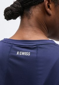 Κοντινό πλάνο από την πλάτη ενός ατόμου με πλεξούδες που φοράει αθλητική μπλούζα K-Swiss σε ναυτικό μπλε χρώμα με ορατές ραφές.